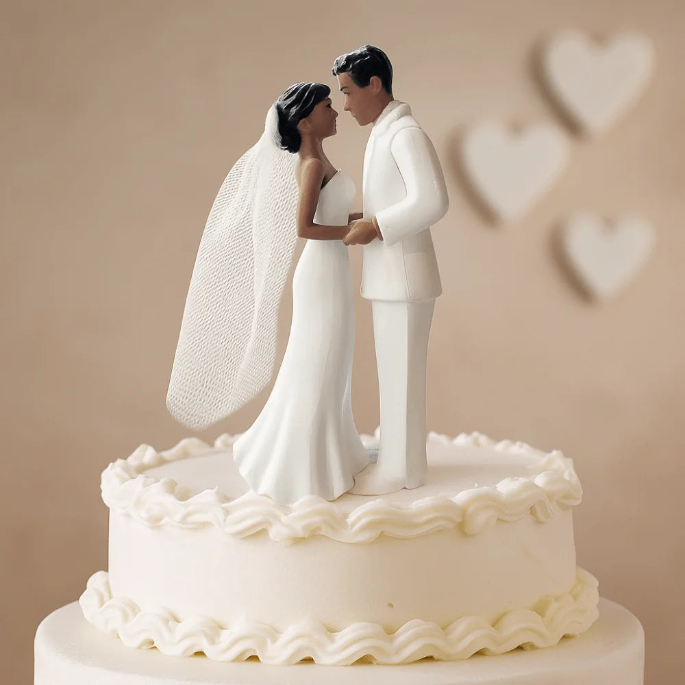 A white cake topper featuring a bride and groom figurine with a veil, set against a light background.