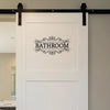 A white sign with the word BATHROOM in black script is mounted on a wall with dark hardware.