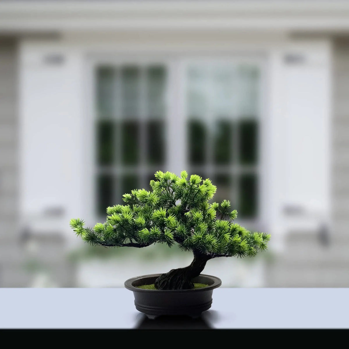 An artificial bonsai tree with green needles is potted in a dark container on a light surface.