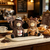 A cute coffee cow figurine with horns and a hat sits on a light tray next to a cup of coffee.