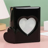 A black candy heart window photocard album with a gold chain detail resting on a pink surface.