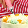 A hand is using a pink piping nozzle to decorate a white cake with orange and mint green frosting swirls.