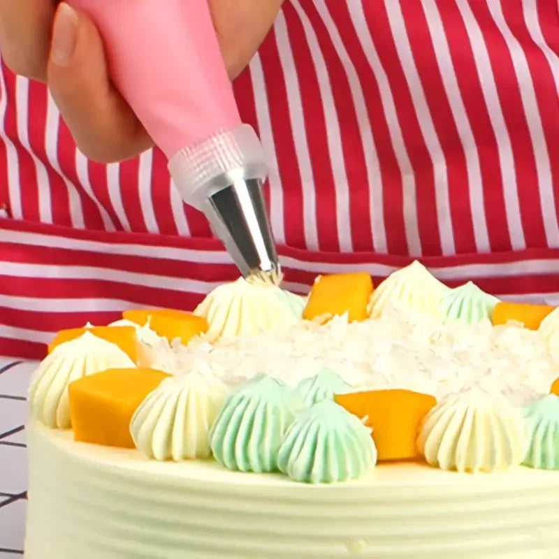 A hand is using a pink piping nozzle to decorate a white cake with orange and mint green frosting swirls.