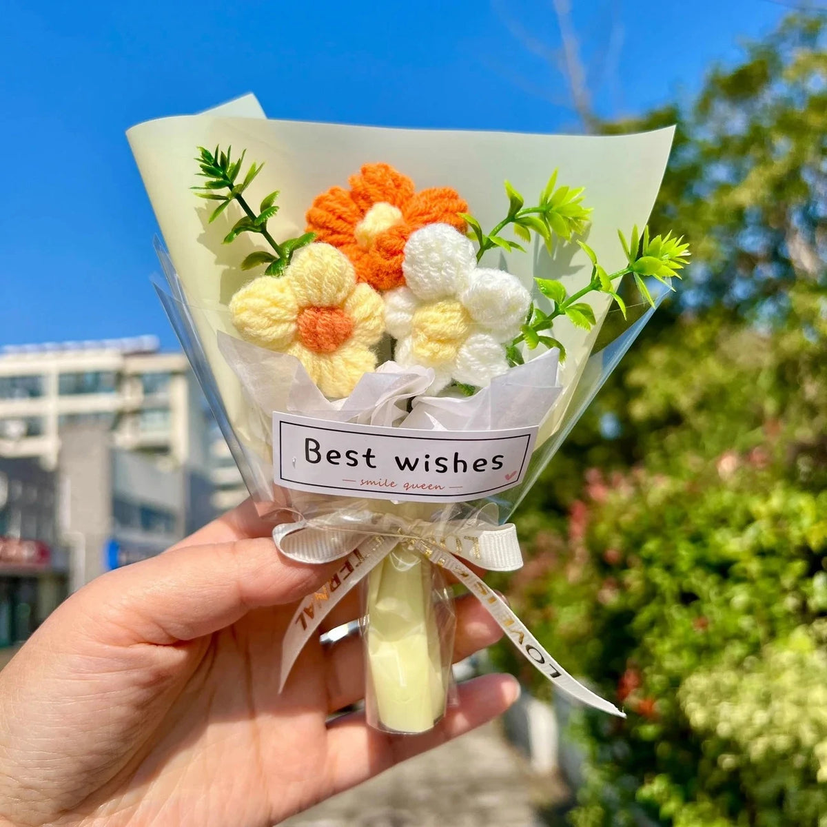 A hand holds a bouquet of handmade crochet flowers in a clear paper wrapping with a white ribbon against a bright blue sky an