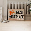 A framed piece of wall art with the text This Must Be The Place on a light background, displayed in a room with a white radia