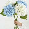 A hand holds a bouquet of blue and white hydrangea silk flower branches with green leaves.