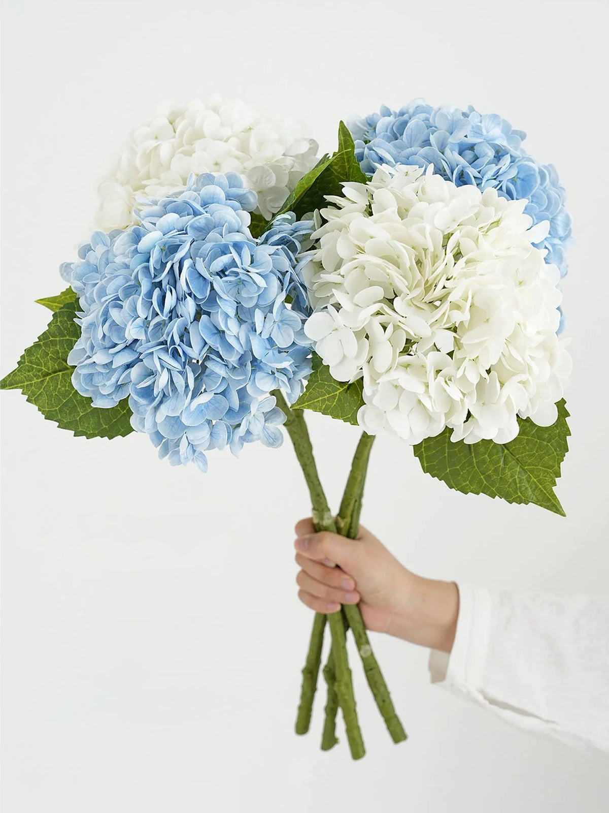 A hand holds a bouquet of blue and white hydrangea silk flower branches with green leaves.