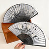 A black and white vintage silk bamboo folding hand fan is shown open, displaying floral patterns.