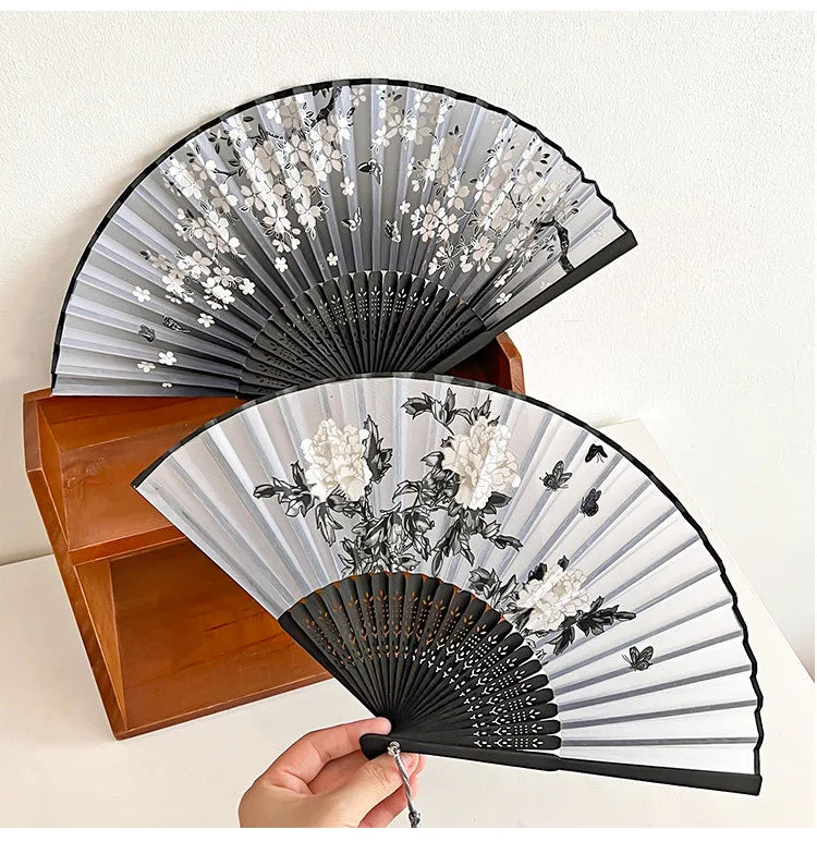 A black and white vintage silk bamboo folding hand fan is shown open, displaying floral patterns.