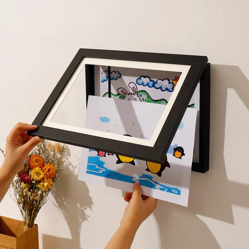 A hand holds a black wooden art display frame with a magnetic closure, featuring colorful children's artwork.