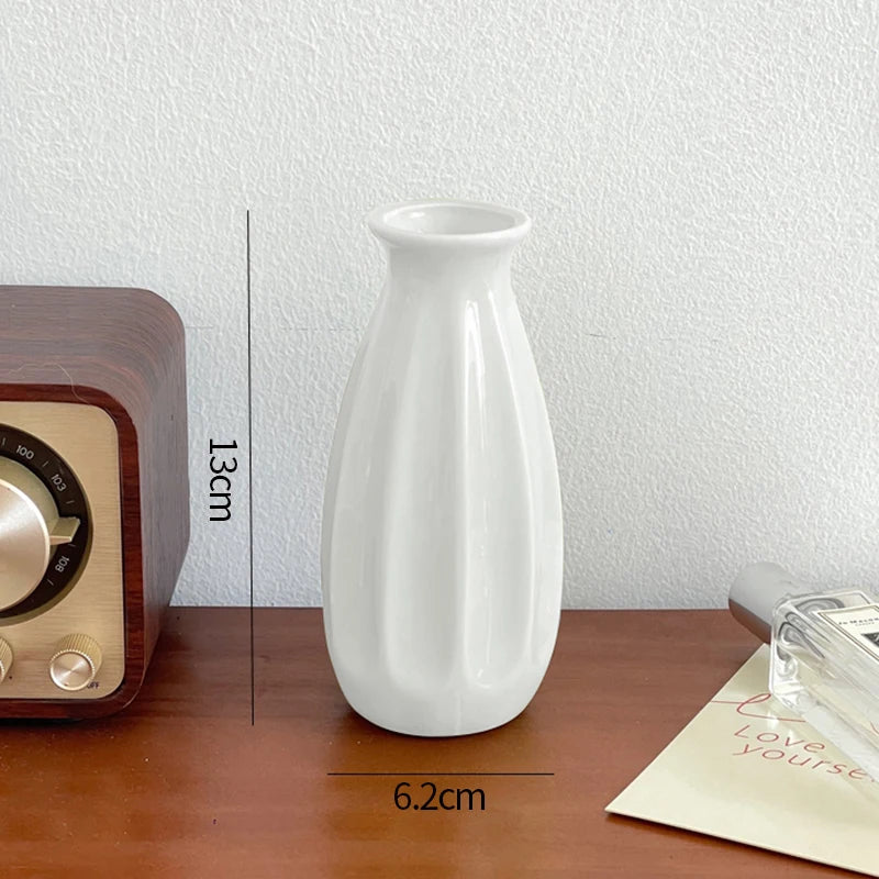 A white ceramic bud vase is shown next to a wooden box and a piece of paper on a brown table.