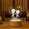 A heart-shaped acrylic night light featuring two cartoon figures and small hearts, resting on a wooden surface.