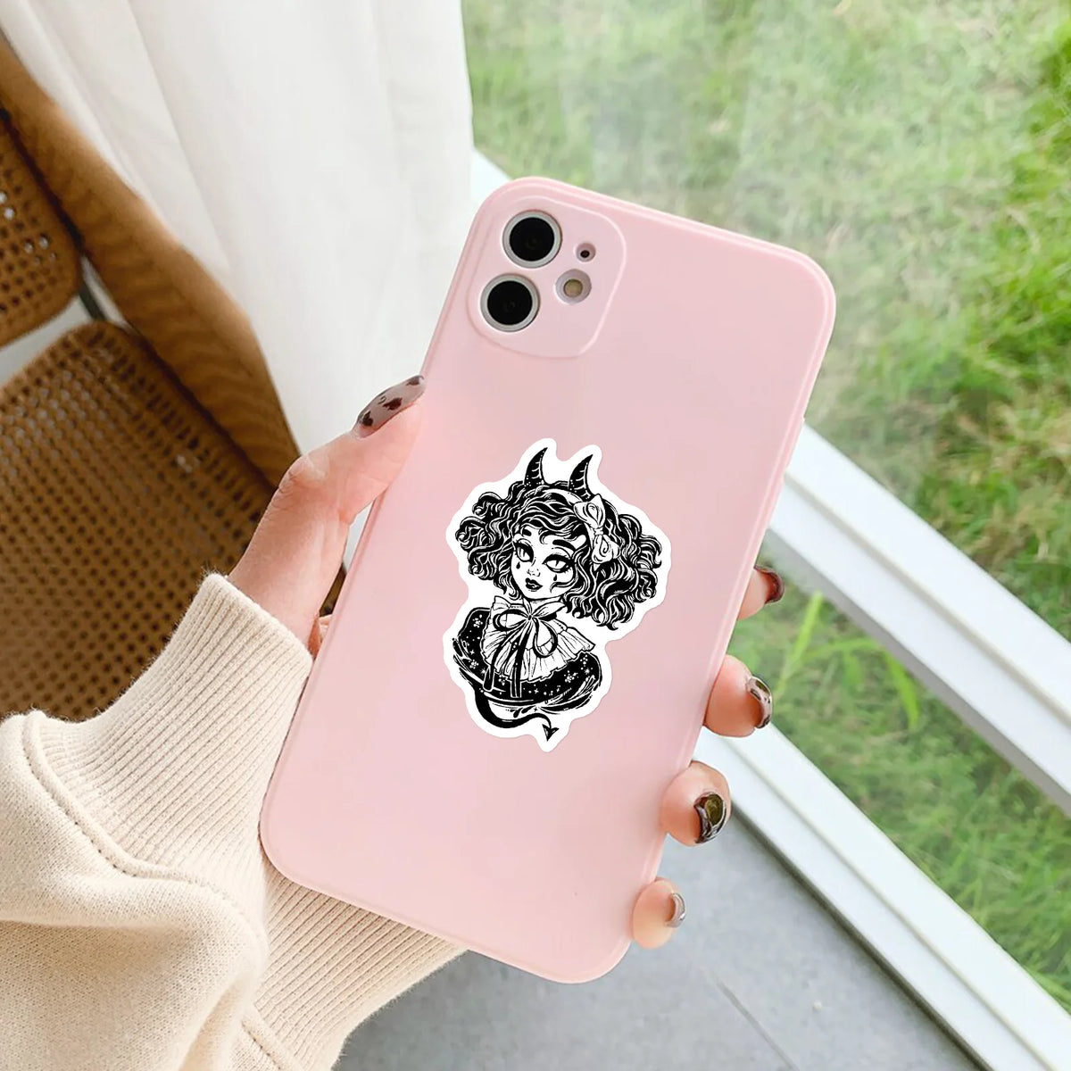 Une main tient un smartphone rose avec un pack d'autocollants de crâne gothique, sur fond extérieur vert flou.