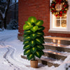 A vibrant green artificial palm tree with 24 leaves stands in a terracotta pot on a snowy path next to a brick house.
