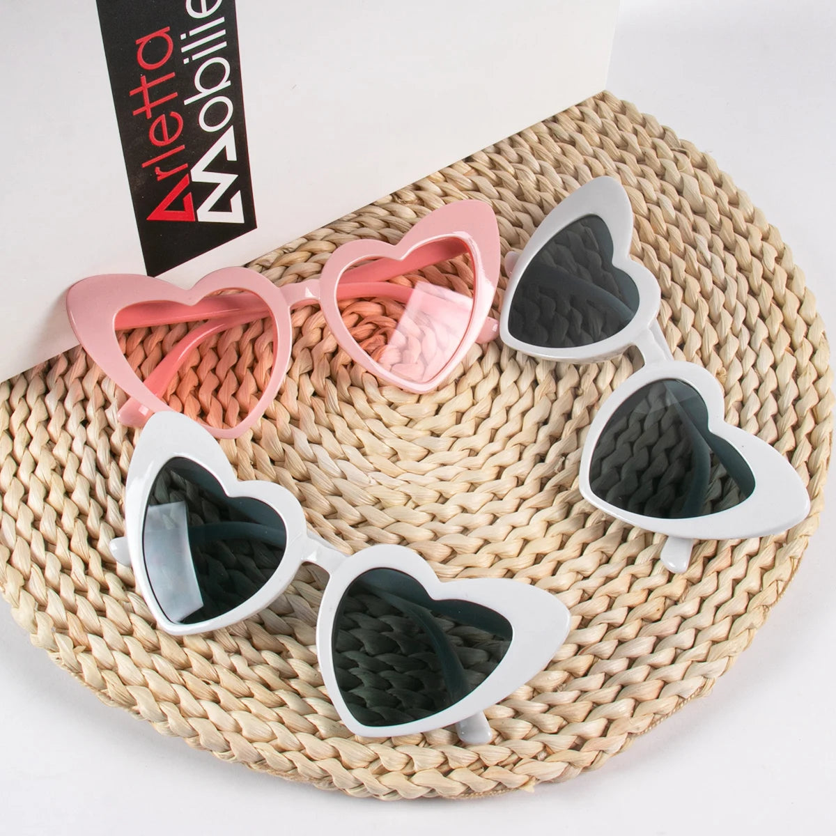 A collection of heart-shaped party sunglasses in pink and silver are displayed on a woven mat.