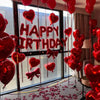 A display of red foil birthday balloons shaped like hearts with bows hanging in front of a window.