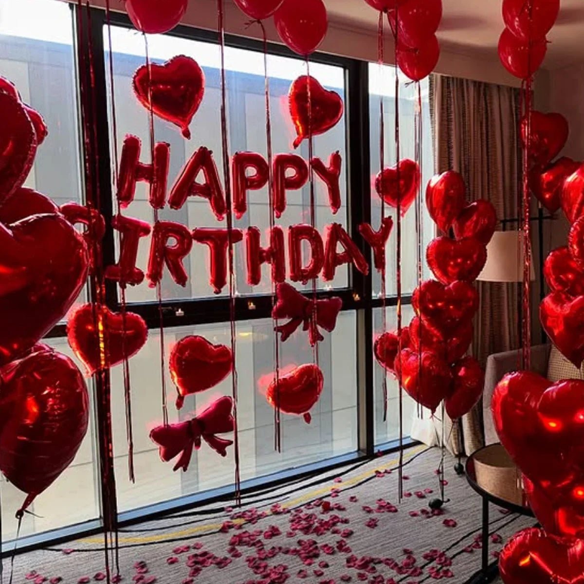 A display of red foil birthday balloons shaped like hearts with bows hanging in front of a window.