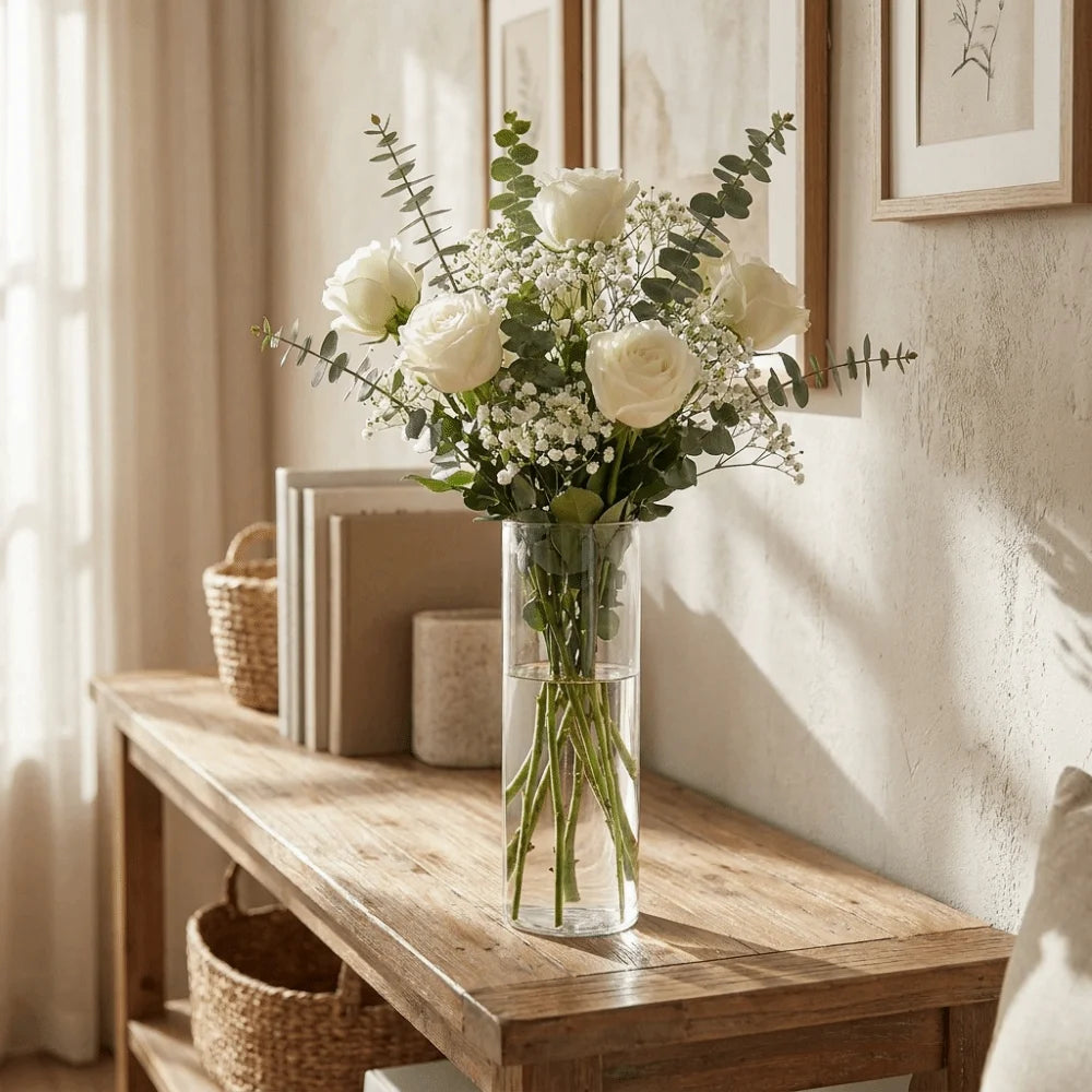 A clear glass cylinder vase filled with white roses and baby's breath flowers on a wooden table.
