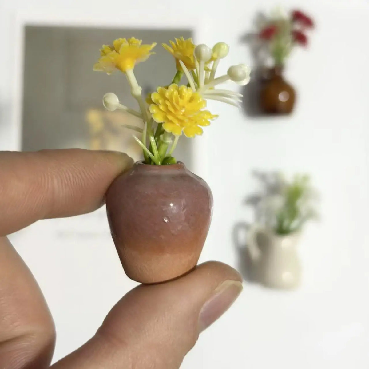 A hand holds a small, earthy-toned ceramic vase with artificial yellow flowers against a white background.