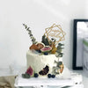 A white cake decorated with green leaves and citrus slices, topped by a gold acrylic birthday sign.