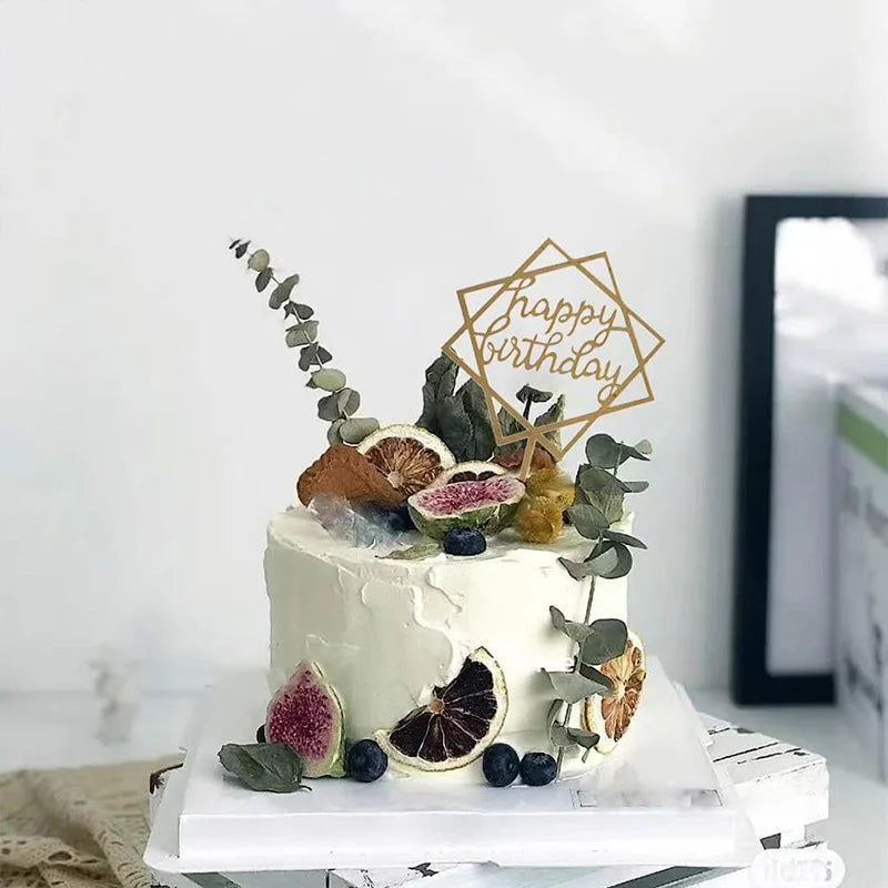 A white cake decorated with green leaves and citrus slices, topped by a gold acrylic birthday sign.