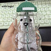 A clear glass coffee cup with a green handle shaped like a bear, held by a hand against a black keyboard.