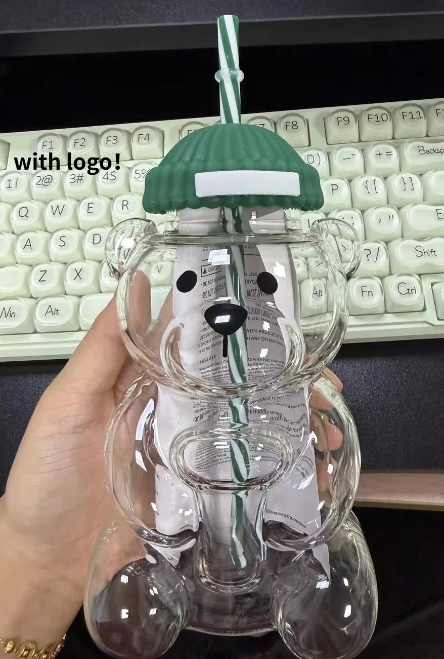 A clear glass coffee cup with a green handle shaped like a bear, held by a hand against a black keyboard.