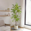 A tall artificial Nandina plant with green leaves in a white pot sits on a windowsill.