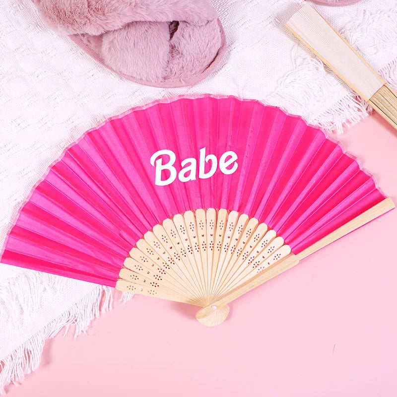 Pink hand fan with the name Babe written on it, resting on a pink surface.
