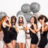 Six silver disco ball foil balloons are held by women in black dresses for party decorations.
