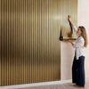 A woman is adjusting a vertical, ribbed gold wallpaper panel on a white wall.