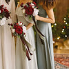 Two women in flowing sage green dresses hold bouquets of artificial red and white roses.