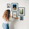A person is hanging five black wooden picture frames on a white wall.