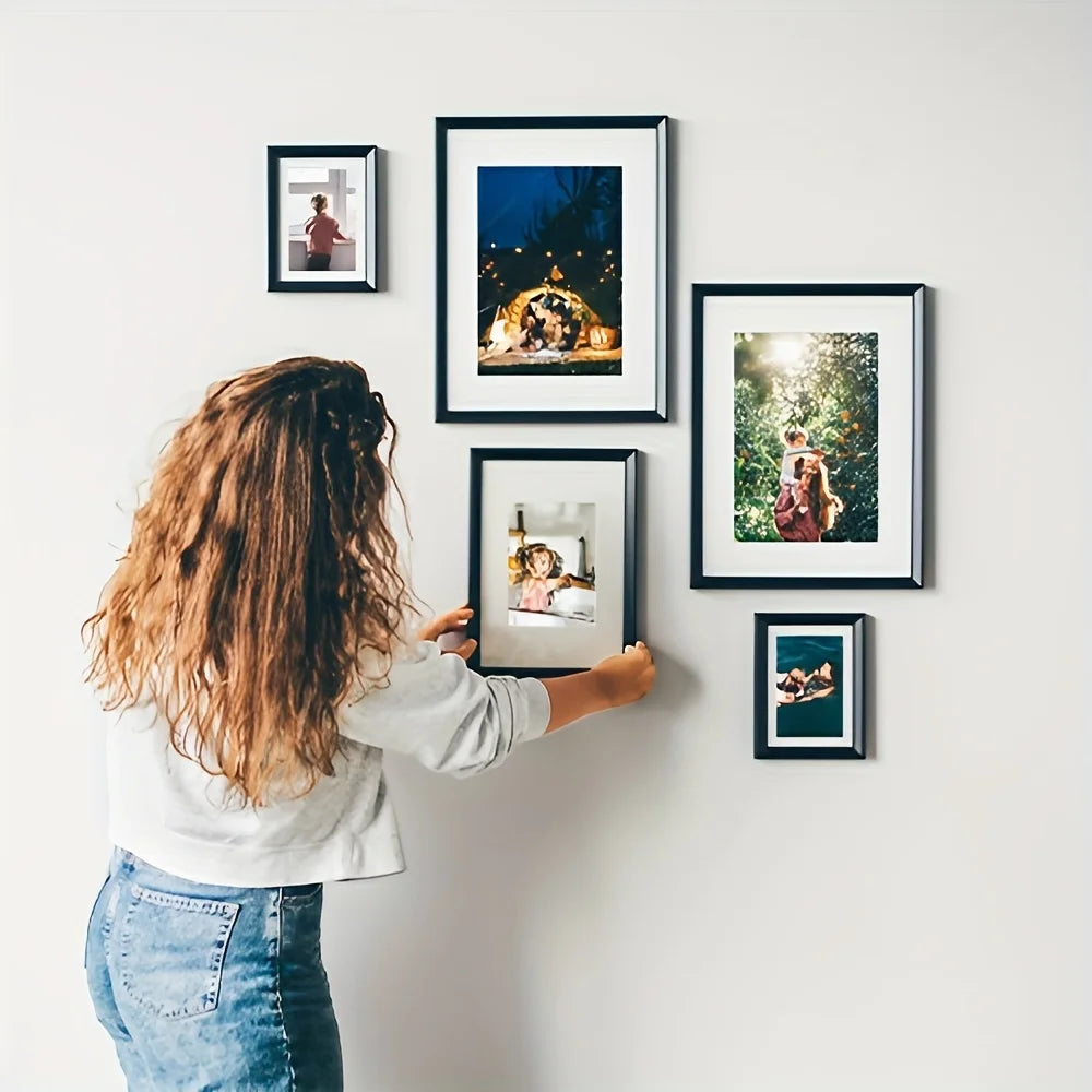 A person is hanging five black wooden picture frames on a white wall.