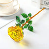 A glass rose flower branch with green leaves and a yellow rose bloom on a white surface.
