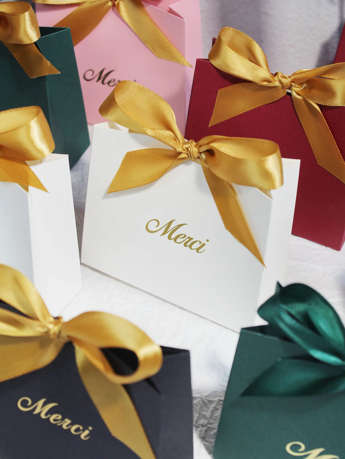 A collection of elegant paperboard thank you gift bags in shades of pink, red, and green with gold ribbon bows.