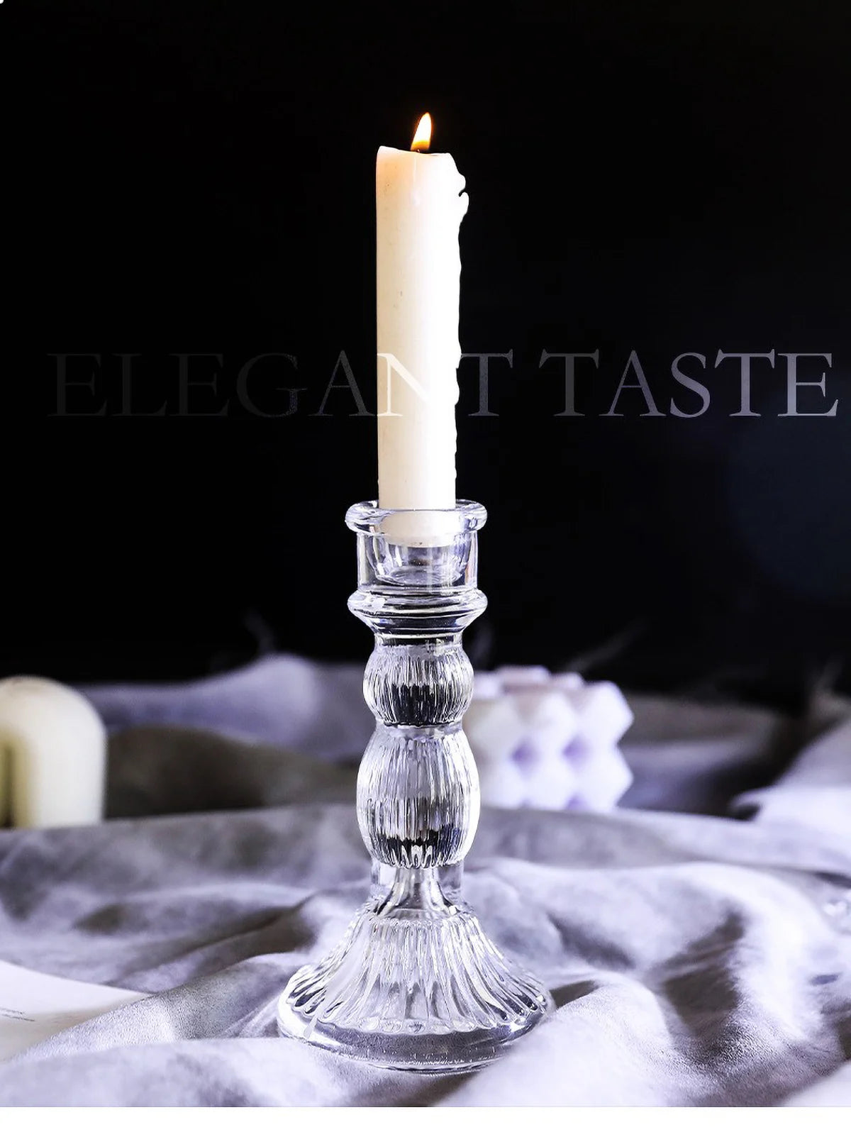 A tall white pillar candle in a clear glass holder resting on a light gray fabric.