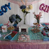 A gender reveal party setup featuring blue and pink foil letter balloons, floral arrangements, and themed treats.