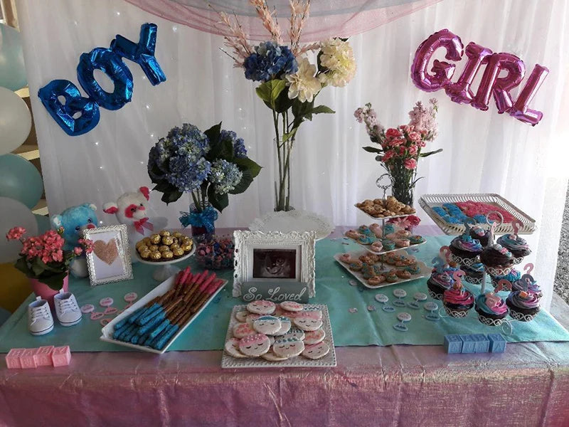 A gender reveal party setup featuring blue and pink foil letter balloons, floral arrangements, and themed treats.