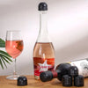 A glass of rosé wine next to a clear glass bottle stopper and a dark silicone wine bottle stopper.