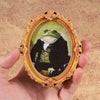 A vintage framed painting of a gentleman frog in a gold ornate frame being held by a hand.