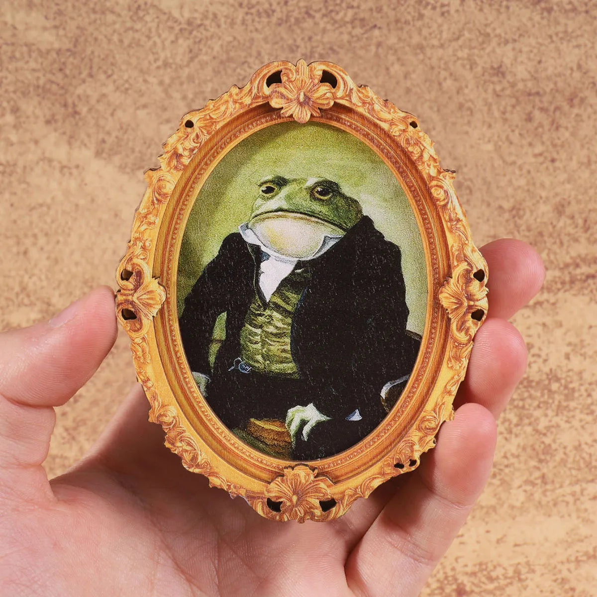 A vintage framed painting of a gentleman frog in a gold ornate frame being held by a hand.