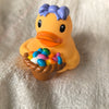 An orange rubber duck toy with blue hair is sitting on a light fabric surface.