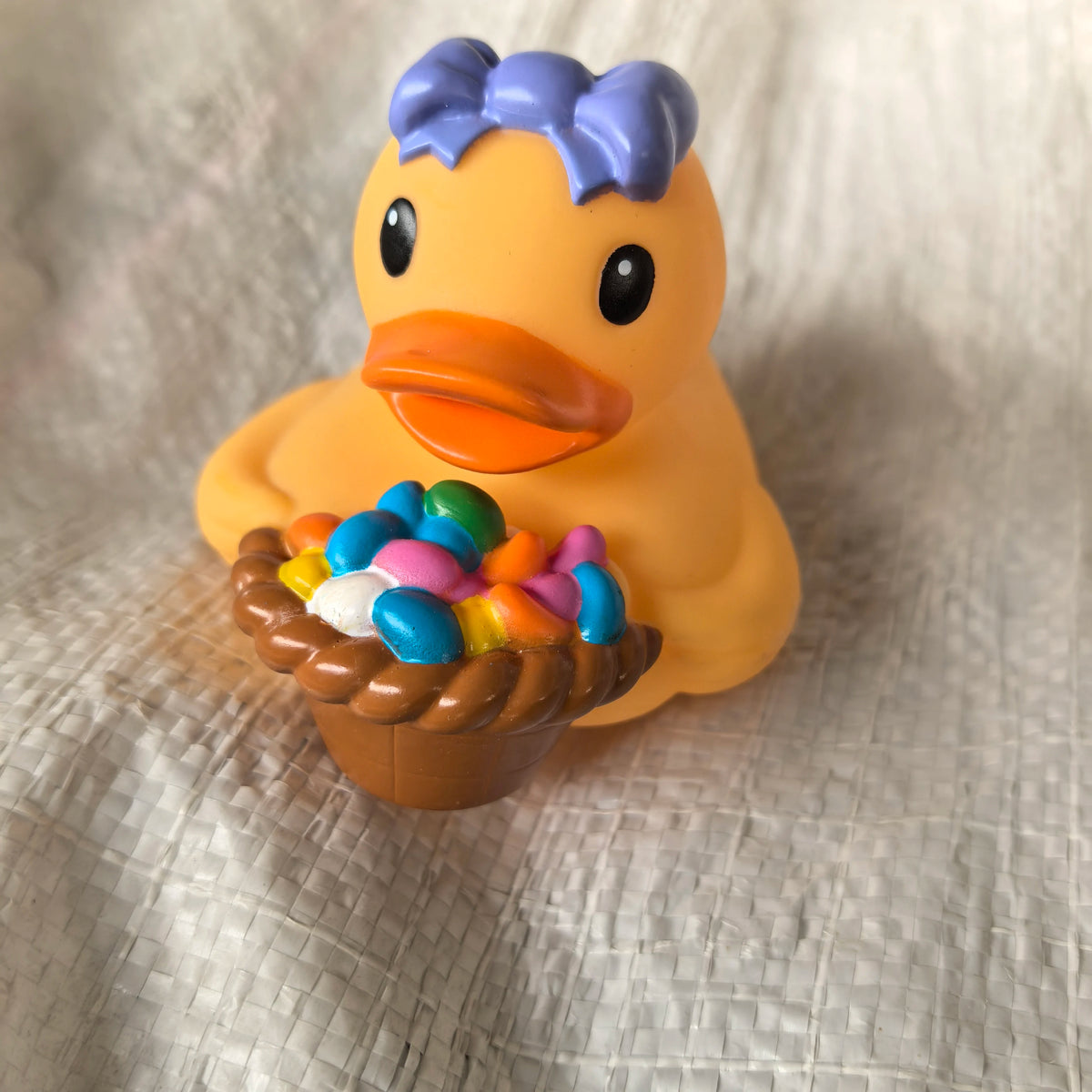 An orange rubber duck toy with blue hair is sitting on a light fabric surface.