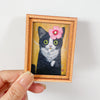 Retrato de acuarela enmarcado de un gato blanco y negro con ojos verdes y flores rosas.