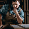 A man wearing an apron is looking down at a wooden surface with papers, possibly working on durable metal tin signs.