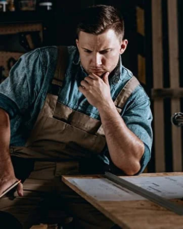 A man wearing an apron is looking down at a wooden surface with papers, possibly working on durable metal tin signs.