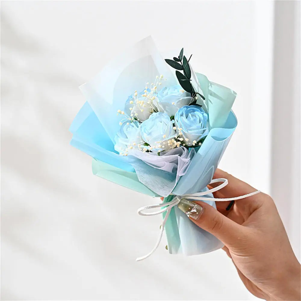 A hand holds a light blue floral bouquet of blue roses and greenery wrapped in sheer fabric.