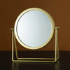 A round, gold-framed makeup mirror with a metal stand, set against a brown background.