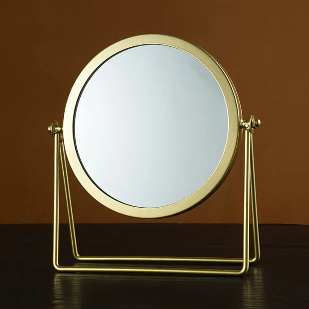 A round, gold-framed makeup mirror with a metal stand, set against a brown background.